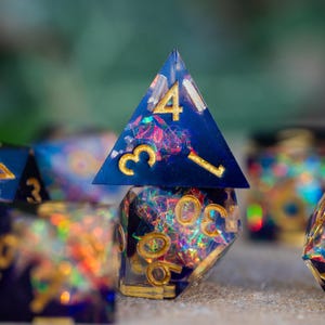 May include: A collection of handmade resin dice in various polyhedral shapes. The dice are a deep blue with gold numbering and colorful inclusions. The image is a close-up, highlighting the intricate details and craftsmanship of the gaming accessories.