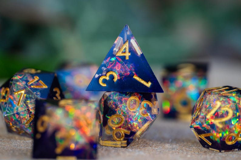 May include: A collection of handmade resin dice in various polyhedral shapes. The dice are a deep blue with gold numbering and colorful inclusions. The image is a close-up, highlighting the intricate details and craftsmanship of the gaming accessories.