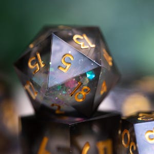 May include: A close-up of a black and gold iridescent d20 dice with glitter embedded in the resin. The dice is showing a 15.
