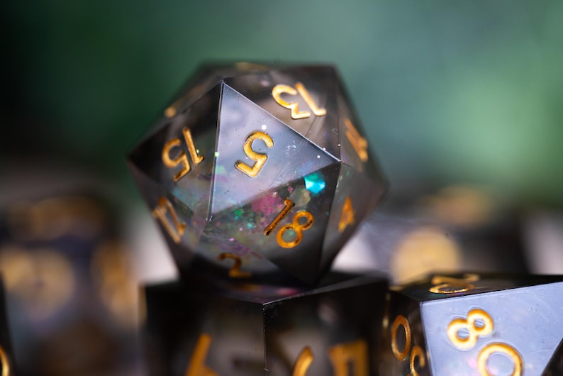 May include: A close-up of a black and gold iridescent d20 dice with glitter embedded in the resin. The dice is showing a 15.