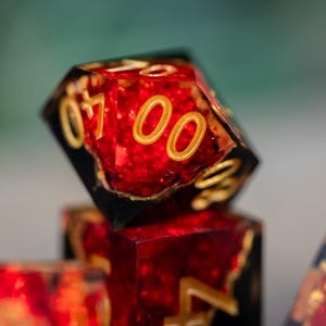 May include: Close-up of a red and black polyhedral dice with gold-colored numbers. The dice has a glittery, textured appearance. The number "0700" is visible on the top face. Other dice are visible in the background.