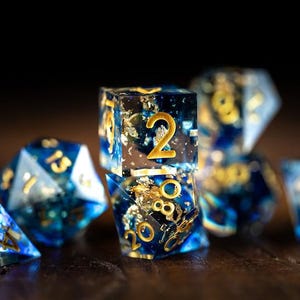 May include: A set of polyhedral dice in blue and gold. The dice are transparent, with gold flakes and gold numbering. The dice are displayed on a dark wooden surface.