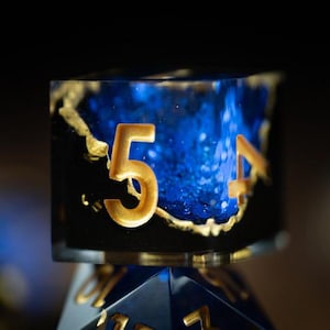 May include: A close-up shot of a blue and black resin die with gold numbers and detailing. The numbers 5 and 4 are visible. The die is set against a dark background.