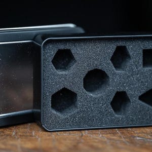 Puede incluir: Caja de dados de metal negro abierta con un inserto de espuma. El inserto tiene recortes para dados de varias formas, incluyendo hex&aacute;gonos, cuadrados y tri&aacute;ngulos. La caja est&aacute; abierta, revelando el interior.