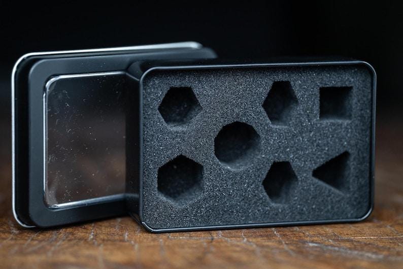 Puede incluir: Caja de dados de metal negro abierta con un inserto de espuma. El inserto tiene recortes para dados de varias formas, incluyendo hex&aacute;gonos, cuadrados y tri&aacute;ngulos. La caja est&aacute; abierta, revelando el interior.