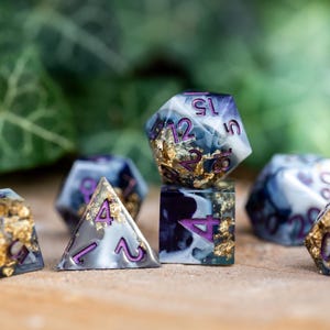 May include: Set of seven polyhedral dice with a white and black marbled design and gold flecks. The dice have purple numbers and are used for tabletop role-playing games.