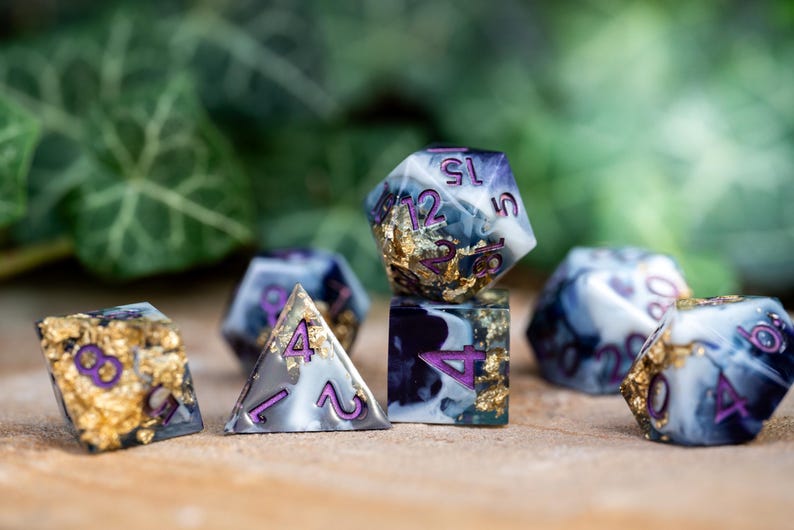 May include: Set of seven polyhedral dice with a white and black marbled design and gold flecks. The dice have purple numbers and are used for tabletop role-playing games.