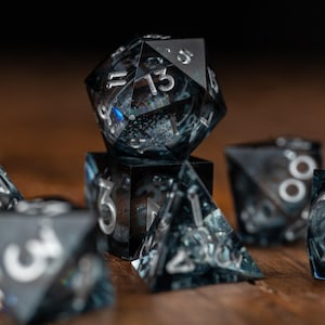 May include: A set of black and clear resin dice with silver numerals. The dice are in various geometric shapes, including a twenty-sided die, a cube, and a pyramid. The dice are on a wooden surface.