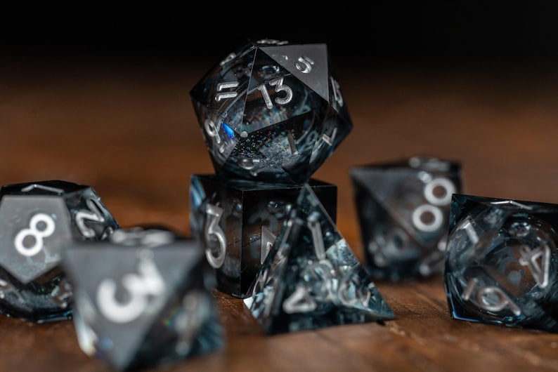 May include: A set of black and clear resin dice with silver numerals. The dice are in various geometric shapes, including a twenty-sided die, a cube, and a pyramid. The dice are on a wooden surface.