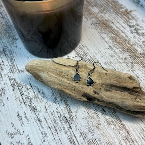 May include: A pair of dangle earrings with a black spade design on a wooden surface. The earrings are made of silver and have a simple design.