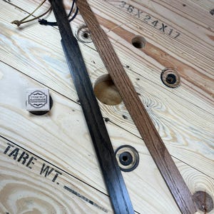 May include: Two wooden paddles, one stained dark brown and the other a lighter brown, are displayed on a wooden surface. The paddles are both about 18 inches long and 2 inches wide. The surface is a wooden spool with the text "36X24X17" and "TARE WT." printed on it.