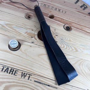 Puede incluir: Un látigo de cuero negro con un mango de madera. El látigo está sobre un carrete de madera con el texto "SERVICE WIRE 36X24X17 TARE WT." impreso en él.