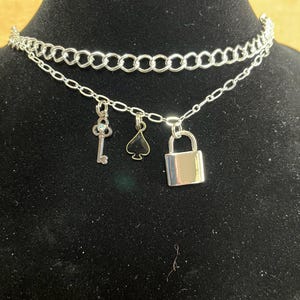 May include: Silver-toned choker necklace with two layers of chain. The top layer is a thicker chain, while the bottom layer has three charms: a key, a spade, and a padlock. The necklace is displayed on a black surface.
