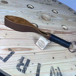 May include: A brown leather strap with a black handle and a wooden end. The strap is on a wooden surface with a stamp that says "Uniquely Handmade, Christian Creations".