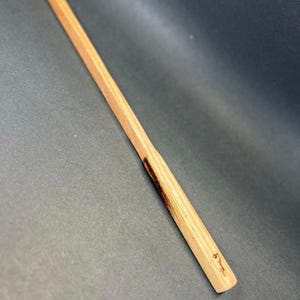 May include: A light-colored wooden wand with a rectangular handle and a tapered body. The wand has a natural wood grain and a small, dark-colored section near the handle. The handle has a small inscription. The wand is set against a dark background.