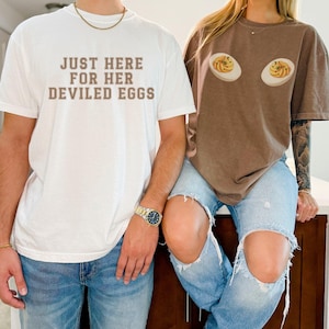 May include: Two people wearing matching t-shirts. One shirt is white with the text "JUST HERE FOR HER DEVILED EGGS" in brown. The other shirt is brown with a graphic of two deviled eggs. Both are wearing blue jeans.
