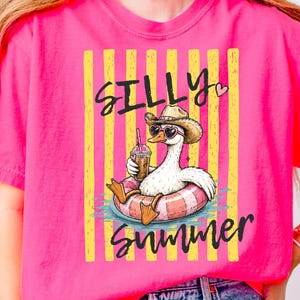 Könnte beinhalten: Ein pinkfarbenes T-Shirt mit einer Cartoon-Gans, die einen Cowboyhut und herzförmige Sonnenbrillen trägt und in einem Pool auf einem rosa-weiß gestreiften Schwimmring schwimmt. Der Text "SILLY SUMMER" befindet sich über und unter der Gans, mit gelben Streifen.