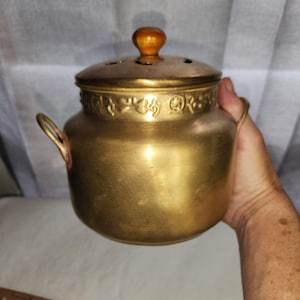 May include: A brass-colored metal pot with two side handles and a removable lid. The lid features a small wooden knob handle and several ventilation holes. A decorative embossed band with repeating patterns encircles the upper neck of the pot.