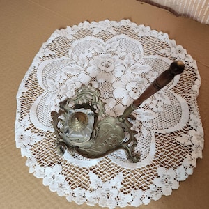 Puede incluir: Un juego de tintero antiguo de latón con un tintero de cristal y una pluma con mango de madera. El tintero tiene un diseño floral ornamentado y se encuentra sobre un tapete de encaje blanco. La pluma está inclinada hacia la derecha.