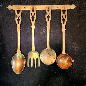 May include: A set of four brass kitchen utensils hanging from a horizontal wall-mounted rack. The set includes a solid spoon, a four-tined fork, a perforated skimmer, and a small ladle, each featuring an ornate, cut-out handle design.