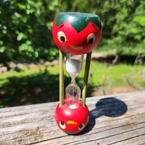 Peut inclure: Un sablier fantaisiste avec un design de tomate rouge et des accents verts. Le centre en verre contient du sable blanc. La structure est soutenue par des tiges métalliques vertes. Un objet décoratif charmant, probablement vintage.