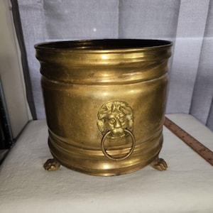 Può includere: Un portavaso cilindrico in ottone caratterizzato da un rilievo a testa di leone con un anello pendente sul lato. Il contenitore poggia su tre piccoli piedini a forma di artiglio ed è posizionato accanto a un righello di legno di circa 20 cm.