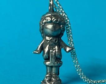 May include: A silver charm necklace featuring a detailed figurine of a person wearing a hat and coat. The charm is attached to a silver chain.