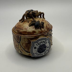 Pode incluir: Um pote de cerâmica redondo com tampa e acabamento manchado em bege e castanho. A tampa apresenta duas aranhas castanhas esculpidas. Um rótulo branco na lateral exibe um gráfico de teia de aranha preta e o texto TRIMMED SPIDER HAIR numa borda decorativa.