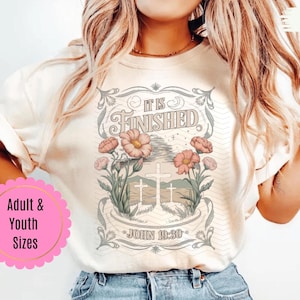 May include: Ivory-colored t-shirt featuring a floral and cross design. The graphic includes the text "IT IS FINISHED" and "JOHN 19:30". The shirt is a relaxed fit and is paired with blue jeans.