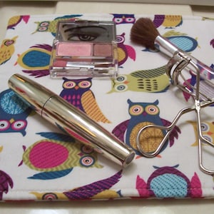 May include: A white makeup bag with a colorful owl print design. The bag contains a silver eyelash curler, a silver mascara wand, a pink and brown eyeshadow palette, and a silver makeup brush.