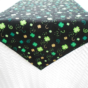 May include: Black table runner with a repeating pattern of green, light green, and gold shamrocks and dots. The shamrocks are in various sizes and are scattered across the fabric. The runner is made of a soft material.