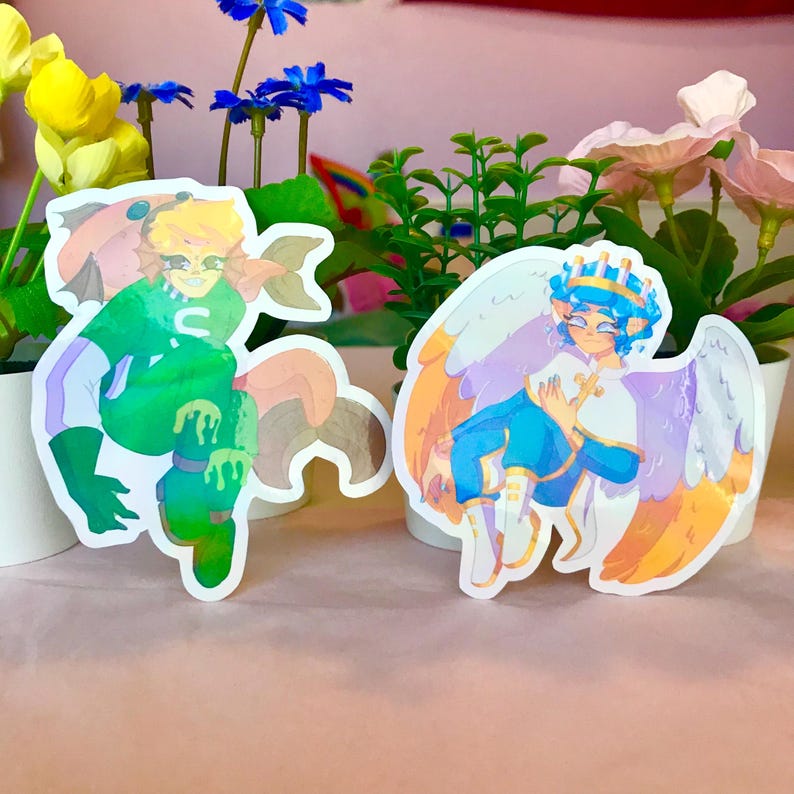 May include: Two holographic stickers featuring cartoon characters. The first sticker is green and features a character with a green and orange fish tail. The second sticker is purple and features a character with a crown and angel wings.
