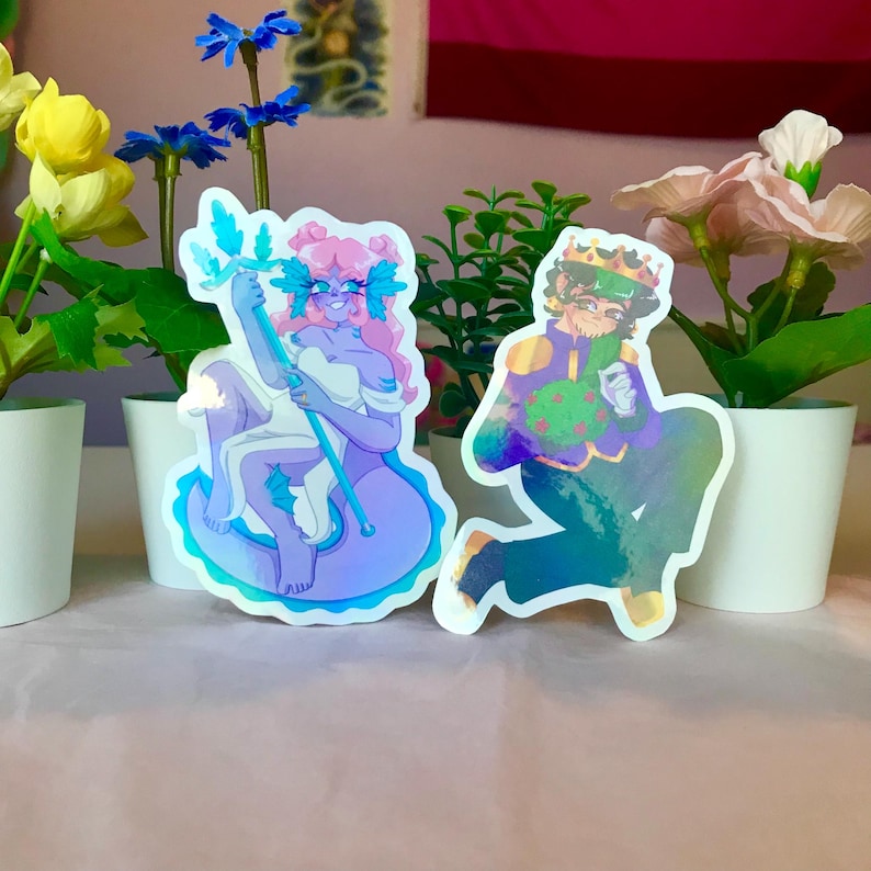 May include: Two iridescent holographic stickers featuring cartoon characters. The first sticker is of a purple-haired woman with a blue tail, holding a blue trident. The second sticker is of a green-haired man wearing a crown and a green and purple outfit.