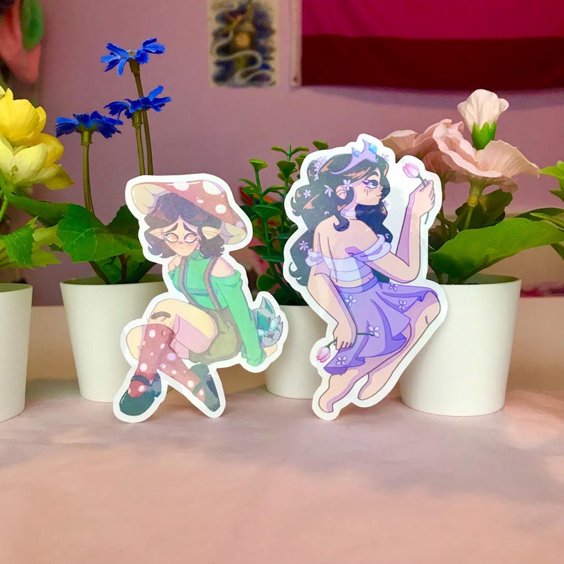 May include: Two holographic stickers featuring illustrations of women. One sticker shows a woman with a mushroom hat and green clothing, sitting with her legs crossed. The other sticker shows a woman with a crown and a purple dress, holding a flower.