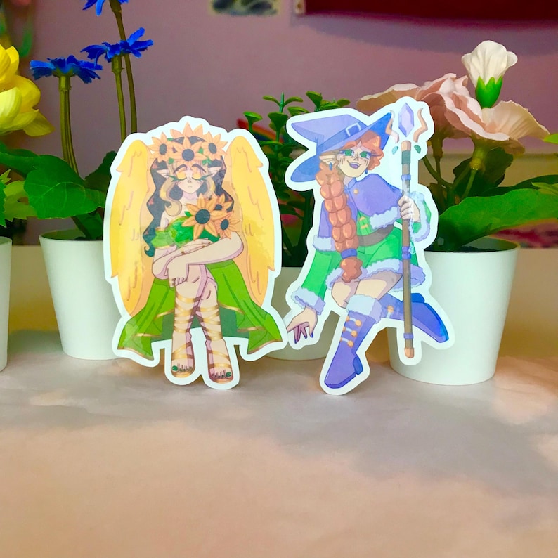 May include: Two illustrated stickers featuring a witch and a sunflower person. The witch is wearing a purple and green outfit with a blue hat and holding a wand. The sunflower person is wearing a green dress with a sunflower crown and has yellow wings.
