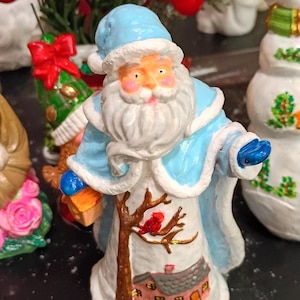 May include: A ceramic Santa Claus figurine in a light blue coat and hat, holding a small gift. The figurine features a detailed design of a house and a bird on a tree. A festive Christmas decoration.