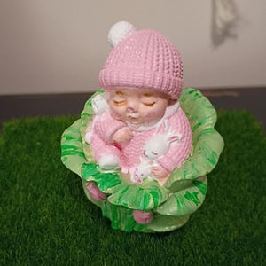 May include: A pink figurine of a sleeping baby wearing a knitted hat and sweater, cradling a white bunny. The baby rests in a green and white cabbage-like structure, set against a green background.