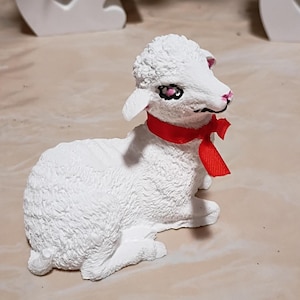 May include: A white lamb figurine with a red ribbon tied around its neck. The lamb has pink accents on its nose and ears, and black and pink eyes. The figurine is sitting down.