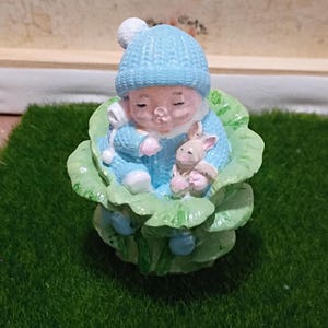 May include: A figurine of a sleeping baby wearing a blue hat and outfit, cradling a small pink bunny. The baby rests in a green leaf-like structure. The word "grandma" is above the figurine.