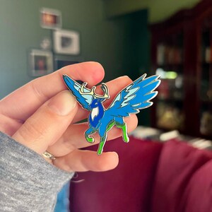 May include: A blue and white enamel pin depicting a winged deer with antlers. The pin features shades of blue, white, and green, with a silver outline. The deer has large, detailed wings and is a fantasy-themed accessory.