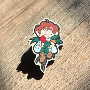 May include: A wooden pin featuring a cartoon character holding a bouquet of flowers and a carrot. The character has brown hair and is wearing a white shirt. The pin is painted in bright colors and has a whimsical style.
