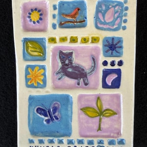 Puede incluir: Una baldosa de cerámica rectangular con fondo crema presenta nueve imágenes cuadradas con varios diseños. Estos incluyen un pájaro, un gato, una mariposa y elementos florales en azul, morado y amarillo. Las palabras "HUMBLE GRACE JOY" están en la parte inferior.