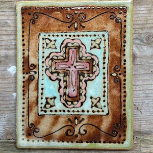May include: A ceramic tile with a brown and green border and a light blue center. The center features a pink cross with a decorative design.