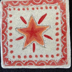 May include: Square ceramic tile with a hand-painted design. The tile features a central star in shades of red and orange, surrounded by a dotted border and a decorative red and white scalloped edge. The background is a speckled off-white.
