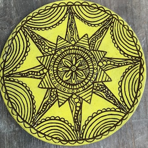 Puede incluir: Un posavasos circular amarillo con un diseño de mandala negro. El mandala presenta una flor central con detalles intrincados y patrones radiantes.