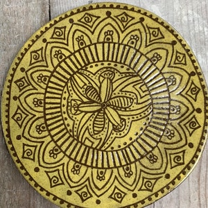 Puede incluir: Un plato de cerámica amarillo con un diseño de mandala marrón. El mandala presenta una flor central con detalles intrincados y un patrón repetitivo de formas geométricas.