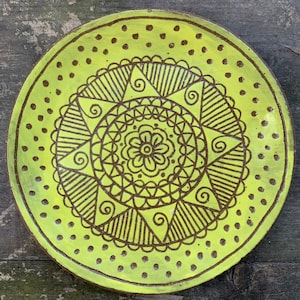 Puede incluir: Un plato redondo de cerámica amarillo con un diseño de mandala negro en el centro. El mandala presenta una flor en el centro rodeada de formas geométricas y espirales.