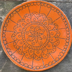 Puede incluir: Un plato redondo de cerámica naranja con un diseño de mandala negro. El mandala presenta una estrella central con patrones en espiral y un diseño floral alrededor del borde exterior.