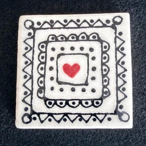 May include: Square ceramic tile with a white background and a black geometric design. The design includes a red heart in the center, surrounded by black dots and lines. The tile has a handmade, folk art aesthetic.