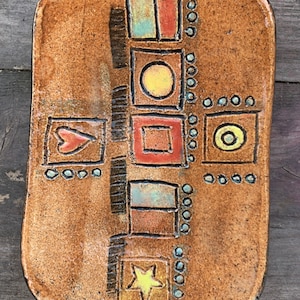 Puede incluir: Un plato rectangular de cerámica con un esmalte marrón y un diseño geométrico en tonos de rojo, amarillo, azul y verde. El diseño presenta círculos, cuadrados y una estrella.